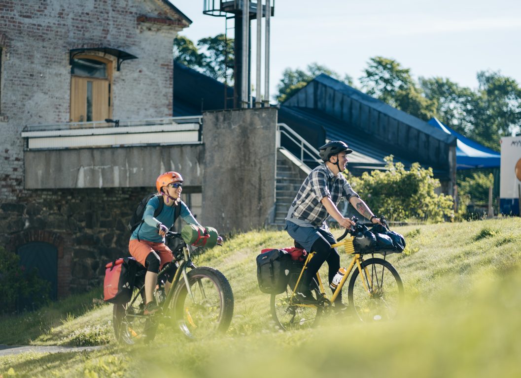 Långfärdscyklister cyklar upp för grön backe i Billnäs med gammal bruksbyggnad i bakgrunden.